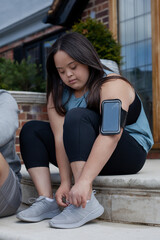 Obraz premium Young biracial woman with Down Syndrome in active wear and wearable tech sitting on steps and tying shoes