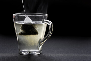 Green tea in a pyramid tea bag is brewed in hot water in a transparent cup. Black background. Back light. Free space for an inscription