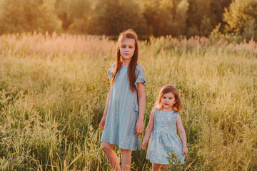 Two little girls wearing blue eco cotton dresses and walking together in summer meadow with green tall grass.