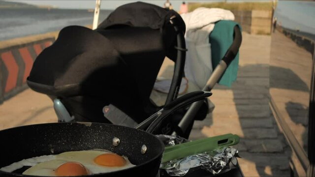 Two eggs are frying in a pan next to a campervan and child's pram on  a sunny day
