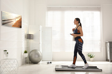 Fototapeta na wymiar Sporty woman with remote control training on walking treadmill and watching TV at home