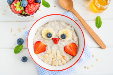 Funny bowl with oat porridge with owl faces made of fruits and berries on a white wooden...
