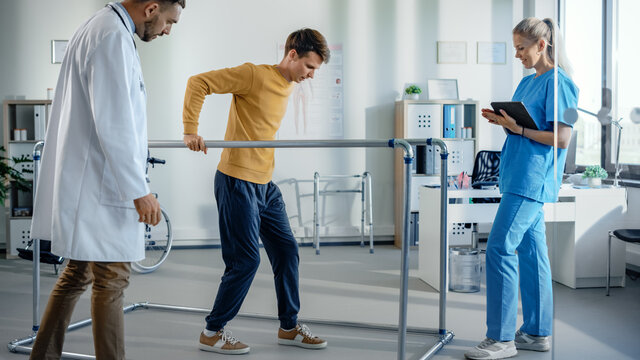 Hospital Physical Therapy Room: Patient with Injury Walking Holding for Parallel Bars, Professional Physiotherapist and Doctor Assist, Help, Train Disabled Person Do Rehabilitative Physiotherapy 