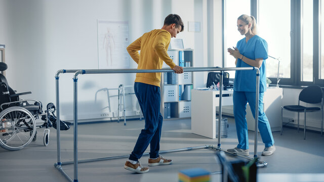 Hospital Physical Therapy Room: Strong Patient with Injury Walking Holding for Parallel Bars, Physiotherapist Encourages Assists, Helps Disabled Person Do Rehabilitative Physiotherapy.