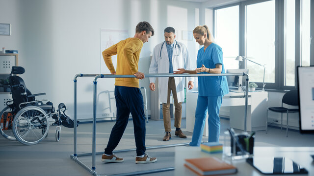 Hospital Physical Therapy: Strong Patient With Injury Making First Steps, Walks Holding For Parallel Bars. Physiotherapist, Rehabilitation Doctor Assist, Help Disabled Person.