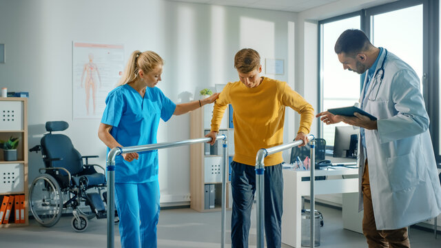 Hospital Physical Therapy: Portrait of Strong Young Male Patient with Injury Successfully Walks Holding Parallel Bars. Physiotherapist, Rehabilitation Doctor, Help, Assist, Disabled Person