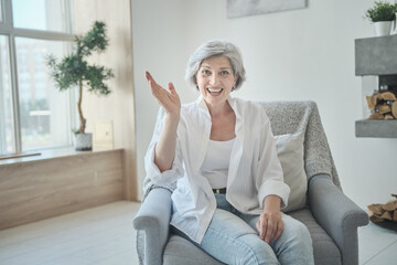 Cheerful elderly woman looking at web camera, enjoying a video call with her daughter and spouse, and an online virtual chat. A modern grandmother in an armchair, making a video call