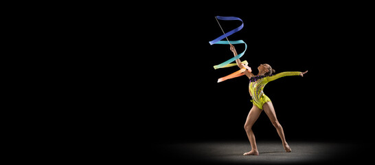 Flyer with little girl, rhythmic gymnastics artist in bright stage costume isolated on dark studio background in ray of light. Concept of sport, action, aspiration, active lifestyle - obrazy, fototapety, plakaty