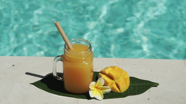 Video footage of glass mason jar with mango juice, bamboo straw, half of fresh mango, yellow frangipani flower and bubbling blue swimming pool on background.
