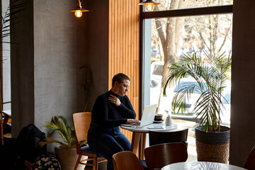 A young girl with short hair and a nose piercing is sitting in a cafe and working on a laptop. The concept of freelancing and remote work or training.