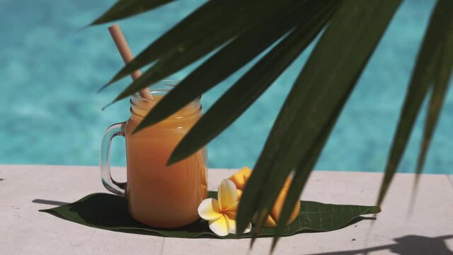 Video footage of glass mason jar with mango juice, bamboo straw, half of fresh mango, yellow frangipani flower, shade from palm tree and bubbling blue swimming pool on background.