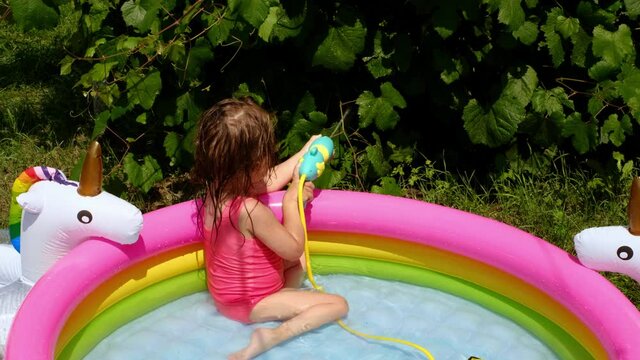 A child is swimming in the pool in the yard and shoots water from a water pistol at a growing grape bush. A water toy gun. Multi-colored inflatable swimming pool for children