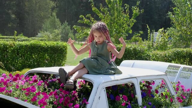 A girl with pink zizi afro - pigtails sits on the roof of a decorative car with flowers growing from inside and plays with her hair, scattering collected pigtails into a bun. Landscape Design Ideas