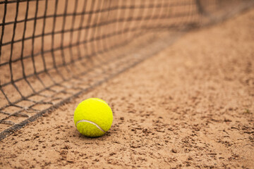 A new tennis ball is lying on a sand court near the net.