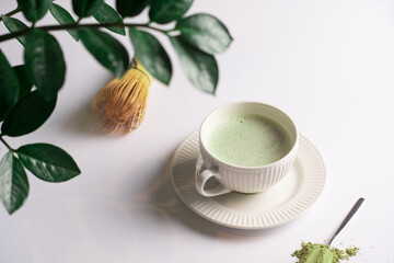 Obraz na płótnie Canvas A white porcelain cup with japanese matcha tea drink on a white saucer plate on a white surface, a tea spoon with matcha powder, bamboos whisker and a green plant zamioculcas