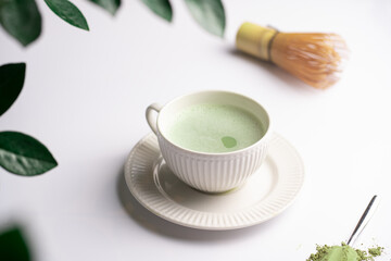 A white porcelain cup with japanese matcha tea drink on a white saucer plate on a white surface, a tea spoon with matcha powder, bamboos whisker and a green plant zamioculcas