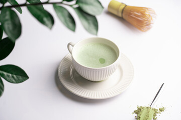 A white porcelain cup with japanese matcha tea drink on a white saucer plate on a white surface, a tea spoon with matcha powder, bamboos whisker and a green plant zamioculcas