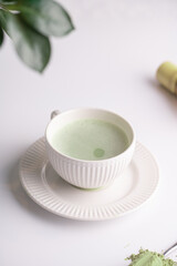 A white porcelain cup with japanese matcha tea drink on a white saucer plate on a white surface, a tea spoon with matcha powder, bamboos whisker and a green plant zamioculcas