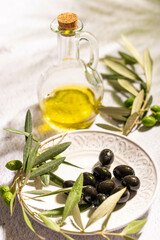 A glass jar with virgin olive oil, textured plate with black olives and a branch of olive tree