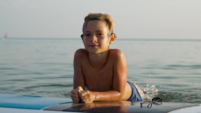 Portrait of child smiling on camera with paddle board on travel holiday at sunset. Happy boy enjoying time in vacation on sea beach.