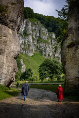 A couple in storm coats walking through a rock gate. 
A picturesque limestone rock formation called "Cracow gate“. 
Ojcow, Polish Jura, Poland.