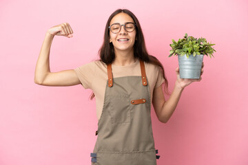 Young gardener girl holding a plant isolated on pink background doing strong gesture