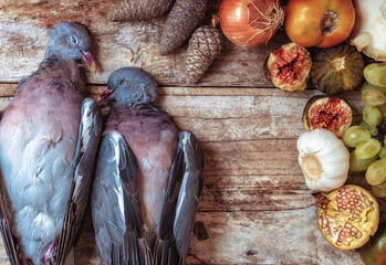 Still life with two wood pigeons on a rustic wooden background, accompanied by fruits and vegetables.