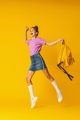 Cute hardworking schoolgirl with a backpack jumps on a yellow background.