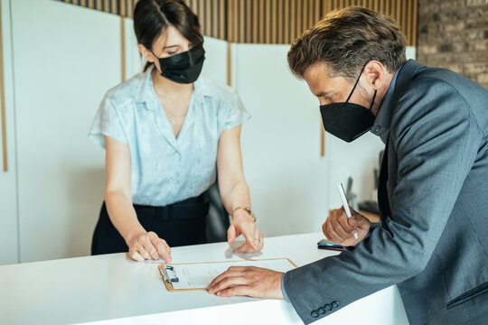 Businessman Checking In At Hotel During COVID-19 Pandemic.
Businessman With KN95 Face Mask Standing At Hotel Reception And Filling In Registration Forms With Assistance Of Female Concierge.