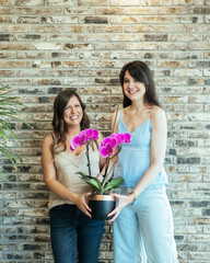 Portrait of Two Smiling Friends Looking at Camera and Holding Potted Orchid Present Together while Standing Against the Brick Wall