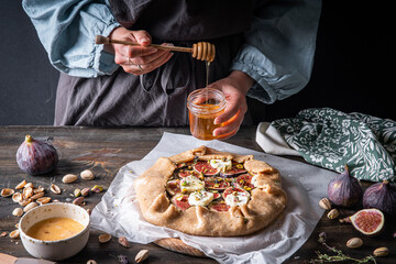Process of making French galette with figs, goat cheese, pistachios and natural honey on baking paper on wooden table.  Woman in rustic closes adding honey.