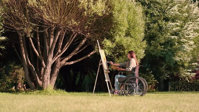 A disabled woman in a wheelchair paints a picture in the park near a tree. Disabled Person, Woman Artist, Art for Sales, Inspiration in Nature.