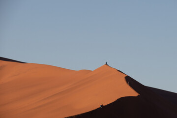 Fototapeta na wymiar Tourists walking and sitting on the edge of a red sand dune at Sossusvlei National Park, a popular tourist destination in Namibia