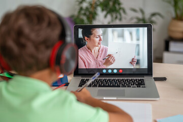 Fototapeta na wymiar little boy with headphones is using laptop to make video call with his teacher. screen displays online lecture with teacher explaining subject from class. E-Education Distance Learning, Home Schooling