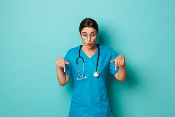 Coronavirus, pandemic and social distancing concept. Image of amazed female doctor in scrubs, pointing and looking down fascinated, standing over blue background