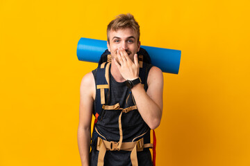 Young mountaineer man with a big backpack isolated on yellow background happy and smiling covering mouth with hand