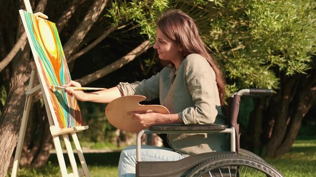 A disabled woman in a wheelchair paints a picture in the park in sunny summer weather. Disabled Person, Woman Artist, Art for Sales, Inspiration in Nature.