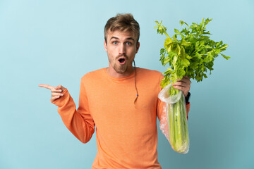 Young blonde man holding a celery isolated on blue background surprised and pointing side