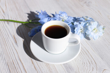 Coffee cup with saucer, blue Delphinium flower on white wood table with shadow. Aroma and good morning concept.