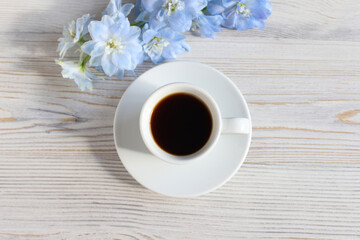Coffee cup with saucer, blue Delphinium flower on white wood table with shadow. Top view, flatlay. Place for your text and product, copy space. Aroma and good morning concept.