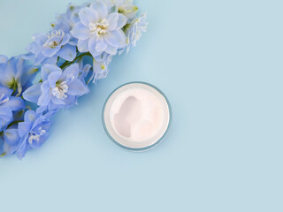 Obraz na płótnie Canvas Moisturizer cream in open glass jar and blue delphinium flower on blue background. Cosmetic bottle blank with hyaluronic acid. Mockup, copy space, top view, flatlay.