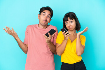 Young mixed race couple holding mobile phone isolated on blue background having doubts while raising hands and shoulders