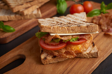 Sandwich with bacon, fried egg. Grilled and pressed toast with bacon, fried egg, tomato and lettuce served on wooden cutting board