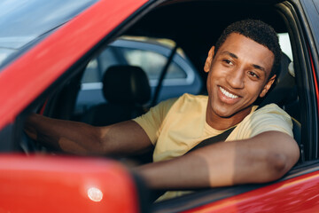 Horizontal view of the young multiracial man smiling while sitting at the car at the driver place and driving car at the traffic jam. Road trip concept