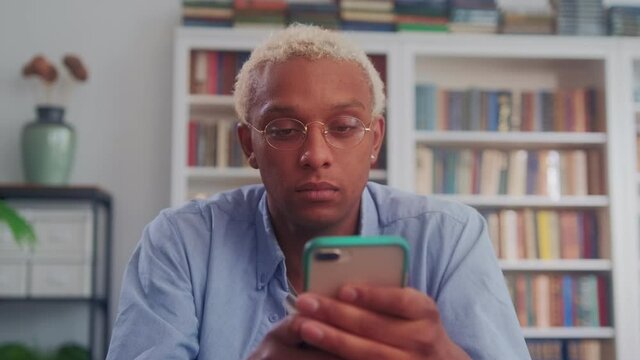 Close up of serious young dark skinned man using smartphone chat with friends sit at desk home office, scrolling feed in aplication. Online communication, business question solution app usage.