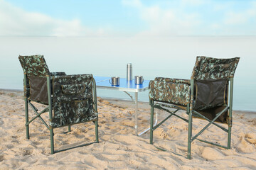 Camouflage fishing chairs and table with metal cups on sandy beach near river