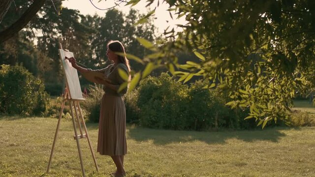 Creative woman artist paints a picture with a brush while standing near the easel in nature. Woman Artist, Art for Sales, Inspiration in Nature.