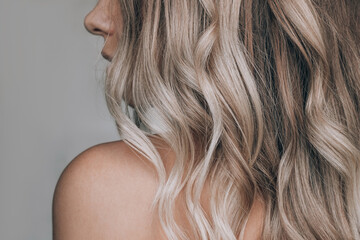 Close-up of the wavy blonde hair of a young blonde woman isolated on a gray background. Result of coloring, highlighting, perming. Beauty and fashion - obrazy, fototapety, plakaty