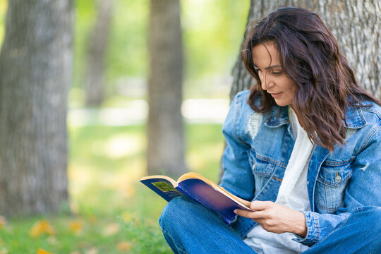 Close-up Portrait Of A 40 Year Old Woman Reading A Book In The Park. She Takes A Break From Working At The Computer And Constantly Online.