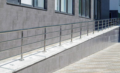 A ramp with stainless steel handrails for wheelchairs, bicycles and strollers with children in front of the building. Handrail for lifting a wheelchair. Shiny stainless steel handrail.